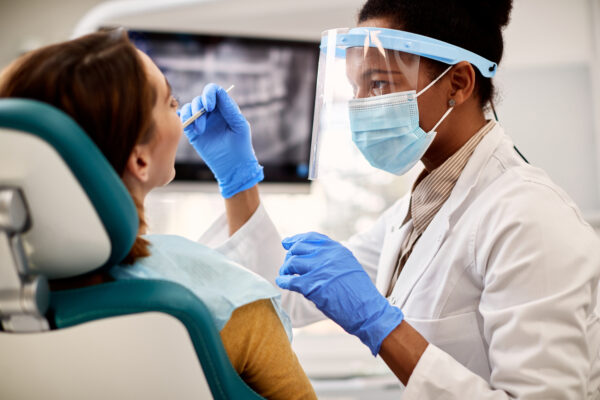 African American dentist with protective workwear performing medical exam on female patient at dental clinic.