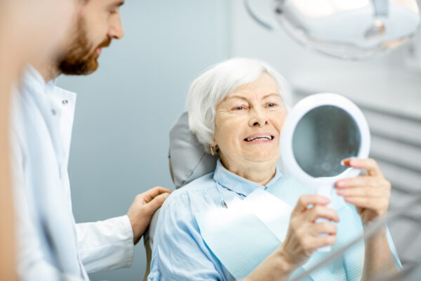 Happy elderly woman enjoying her beautiful toothy smile looking to the mirror in the dental office