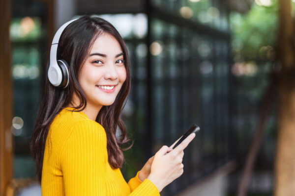 Happiness Asian smiling young woman wearing technology wireless headphones for listening the music via smart mobile phone infront of coffee shop or coworking space, Lifestyle and leisure concept