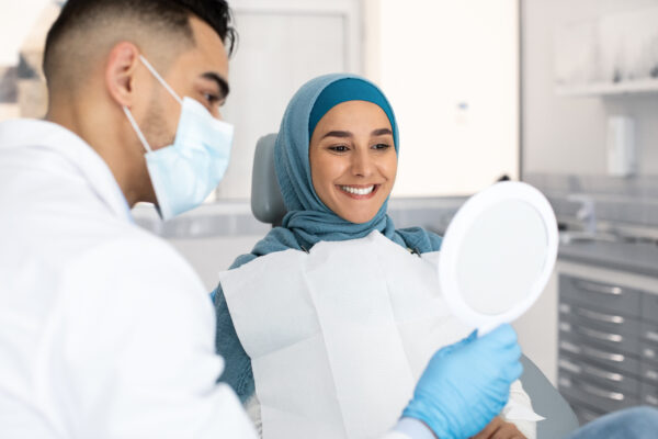 Muslim Lady In Hijab Looking At Her New Smile After Teeth Treatment In Dental Clinic, Middle Eastern Dentist Holding Mirror, Showing Result Of Medical Procedures For Islamic Female Patient, Closeup