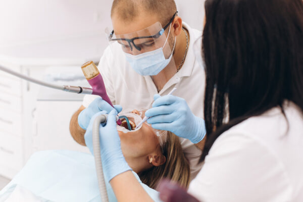 A dentist in a mask and goggles treats a woman's teeth.