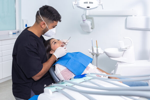 Male dentist in gloves and face mask using mirror to inspect teeth of teen patient during check up in light office of dental clinic